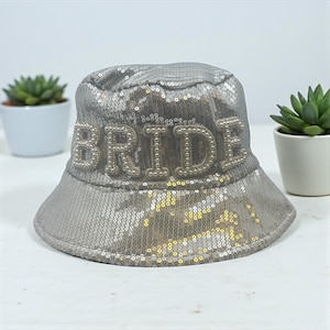 May include: A silver sequined bucket hat with the word "BRIDE" in pearl beads. The hat is a light silver colour and has a wide brim. A bridal accessory.