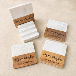 May include: A set of square coasters featuring a white marble top and a brown wood base. Each coaster is engraved with the text Oli & Angélica and Est: 10th Sept 2026. A matching marble and wood holder is included to store the coasters vertically.