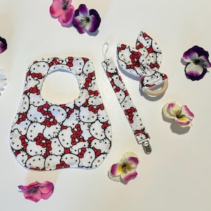 May include: A white bib with a red and white Hello Kitty print, a white and red Hello Kitty clip-on teether, and a wooden teether with a white and red Hello Kitty print.