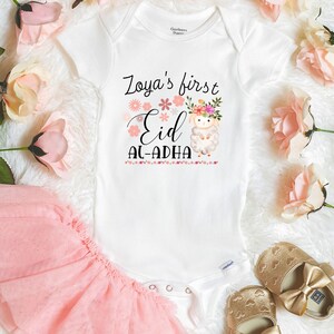 May include: White onesie with a pink floral design and the text "Zoya's first Eid Al-Adha". A white lamb with a pink floral crown is in the center of the design.