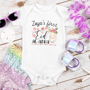 May include: White baby onesie with a pink floral design and a cute lamb. The onesie says "Zoya's first Eid Al-Adha".