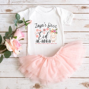 May include: White onesie with a pink floral design and the text "Zoya's first Eid Al-Adha". A white lamb with a floral crown is in the center of the design. The onesie is paired with a pink tulle skirt.