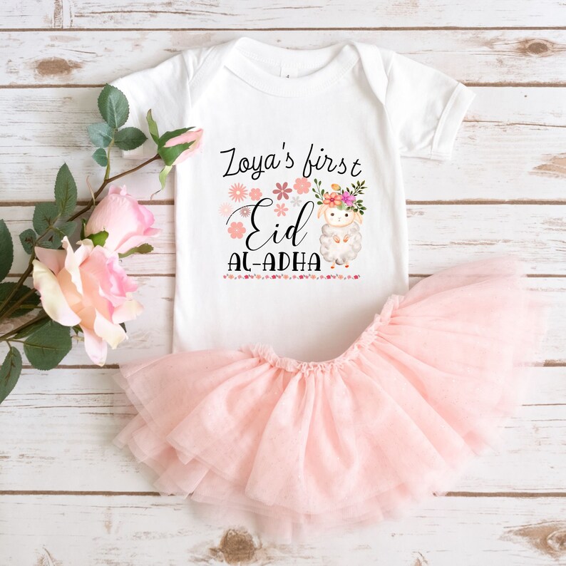 May include: White onesie with a pink floral design and the text "Zoya's first Eid Al-Adha". A white lamb with a floral crown is in the center of the design. The onesie is paired with a pink tulle skirt.