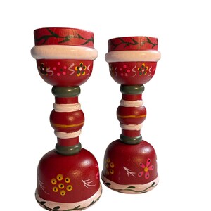 May include: Two red wooden candle holders with white, green, and yellow painted designs. The candle holders have a bell-shaped base and a cylindrical middle section.
