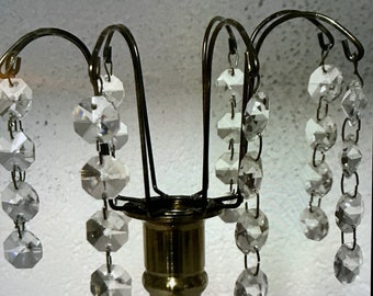 Peut inclure: Un bougeoir de couleur laiton avec un design décoratif. Le bougeoir présente des bras métalliques incurvés d'où pendent des perles de cristal claires et facettées. La base est de forme classique.