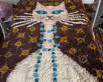 May include: A white cat with blue eyes and a turquoise necklace is depicted on a brown and gold patterned rug. The rug is made of a fluffy, textured material.
