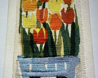 May include: A woven wall hanging featuring a bouquet of colorful tulips in a blue pot. The tulips are red, orange, yellow, and green. The pot has a white and blue pattern.
