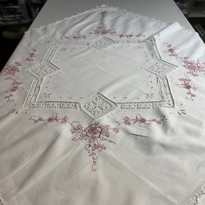 Puede incluir: Mantel cuadrado blanco con un diseño floral en color rosa apagado. El mantel presenta flores bordadas, inserciones de encaje geométrico y un borde de ganchillo. Textil de estilo vintage.