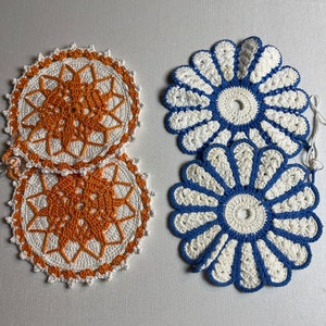Puede incluir: Dos juegos de posavasos de ganchillo. Un juego es blanco y naranja, el otro es blanco y azul.