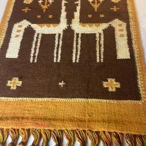 May include: A rectangular brown woven textile with a repeating pattern of two stylized white llamas. The textile has a gold border and a fringe of knotted tassels in shades of gold, orange, and brown. Decorative crosses and floral motifs are also present.