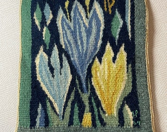 May include: A square, woven textile featuring a floral design. The central motif showcases three stylized flowers in shades of blue and yellow against a dark blue background. The border is green, and the edges are finished with a gold trim.