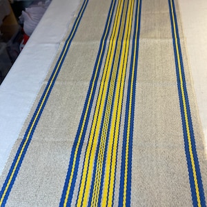 Puede incluir: Un camino de mesa tejido beige con rayas verticales azules y amarillas. El camino de mesa tiene un borde con flecos en ambos extremos. Las rayas están espaciadas uniformemente y recorren toda la longitud del camino.