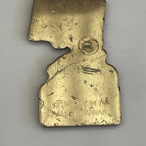 May include: A vintage brass-colored metal pin in the shape of a state. The pin has a worn, textured surface with visible scratches. The text "&copy;1991 WC 94" and "MADE IN TAIWAN" are stamped on the pin.