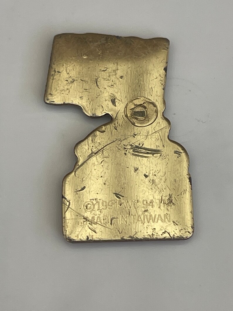 May include: A vintage brass-colored metal pin in the shape of a state. The pin has a worn, textured surface with visible scratches. The text "&copy;1991 WC 94" and "MADE IN TAIWAN" are stamped on the pin.