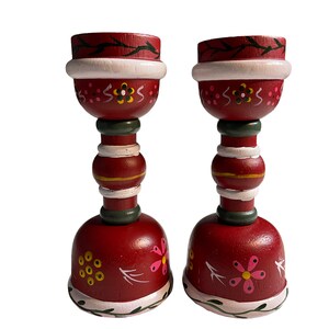 May include: A pair of red wooden candle holders with white, green, and pink painted floral designs. The candle holders have a bell-shaped base and a cylindrical middle section.