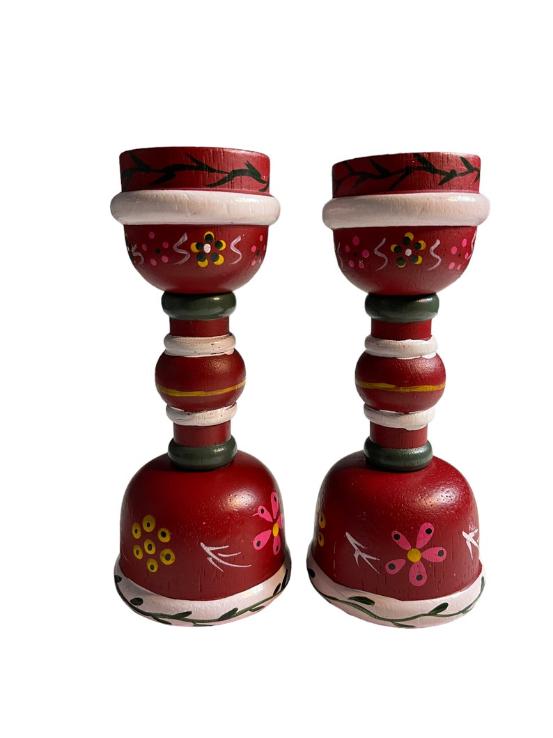 May include: A pair of red wooden candle holders with white, green, and pink painted floral designs. The candle holders have a bell-shaped base and a cylindrical middle section.