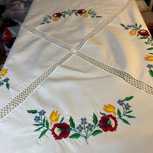 Könnte beinhalten: Weiße Tischdecke mit gestickten Blumendesigns in Rot, Gelb und Blau. Die Tischdecke hat einen Häkelrand und ein Kreuzstichdesign in der Mitte.