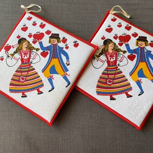 Op de afbeelding: Twee witte pottenlapjes met rode bies tonen een volkskunstontwerp van twee paren in traditionele kleding die rode harten vasthouden. De achtergrond is gevuld met rode harten.