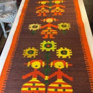 Puede incluir: Un corredor tejido marrón con un borde naranja. El corredor presenta un patrón repetido de figuras estilizadas en naranja, amarillo y verde, tomados de la mano y de pie junto a grandes flores amarillas. El diseño es simétrico e inspirado en el arte popular.