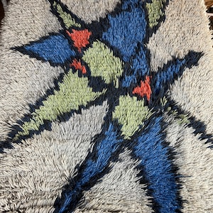 May include: A vintage rug with a geometric design featuring a large, stylized star shape in shades of blue, green, black, and orange. The star is set against a light gray background.