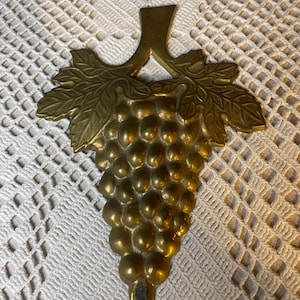 May include: A brass grape cluster wall hanging. The decorative piece features a detailed design of a bunch of grapes with leaves and a stem. The brass has a shiny, golden finish, and the grapes are round and textured.