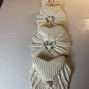 May include: A cream-colored macrame plant hanger with four woven pockets. The pockets are circular and connected by a vertical rope. The macrame is made of thick, off-white cotton cord, and the top has a wooden ring for hanging.