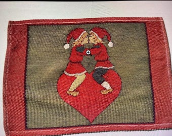 May include: A rectangular tapestry with a red border and a central design featuring two figures in red outfits and hats kissing atop a red heart. The background is olive green.