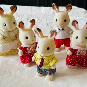 May include: A family of five white rabbit figurines with brown ears. The mother rabbit is wearing a yellow dress with a white apron and lace trim. The father rabbit is wearing a white shirt with a red bow tie and red pants. The three children are wearing red dresses with white lace trim and white polka dots.