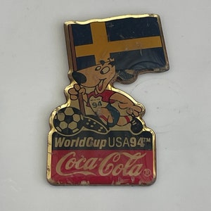 May include: A vintage pin featuring a cartoon dog with a soccer ball, holding a flag of Sweden. The pin is for the 1994 World Cup USA, with the Coca-Cola logo at the bottom. The pin has a gold border.