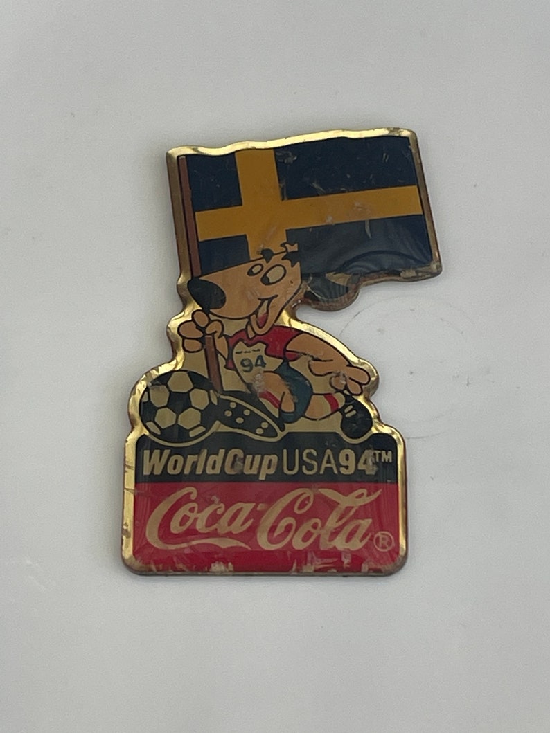 May include: A vintage pin featuring a cartoon dog with a soccer ball, holding a flag of Sweden. The pin is for the 1994 World Cup USA, with the Coca-Cola logo at the bottom. The pin has a gold border.
