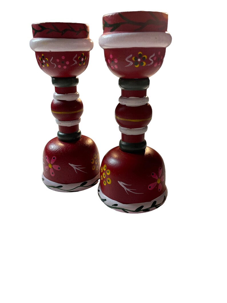 May include: A set of two red wooden candle holders with white and green painted details. The holders are decorated with pink and yellow flowers and have a traditional folk art style.