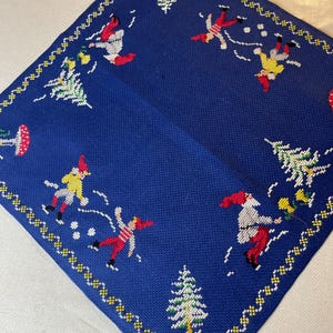 May include: A square, blue embroidered cloth featuring a repeating pattern of skiers and Christmas trees. The skiers are depicted in red, yellow, and white outfits. The border is a repeating pattern of yellow and white stitches.