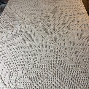 May include: A cream-colored, handmade crochet blanket. The blanket features a detailed, openwork design with geometric and floral patterns. The texture is visible, showcasing the intricate craftsmanship. The blanket is spread out, suggesting it's a bedspread or decorative throw.