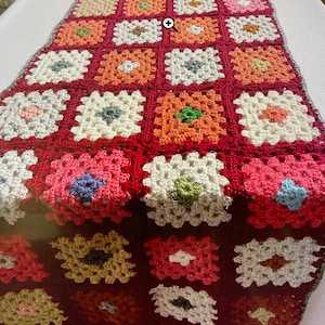 Puede incluir: Un camino de mesa tejido a crochet con un patrón de cuadrados de la abuela en tonos de rojo, blanco, naranja, rosa y verde.