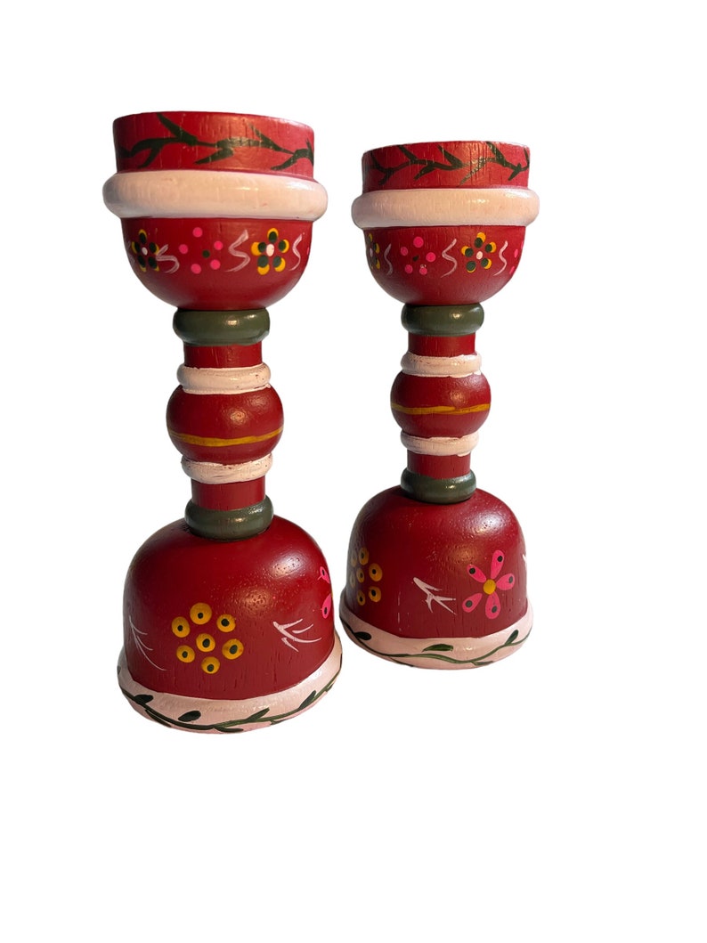 May include: Two red wooden candle holders with white, green, and yellow painted designs. The candle holders have a bell-shaped base and a cylindrical middle section.