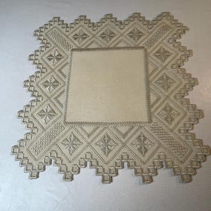 Peut inclure: Une nappe carrée en lin blanc cassé avec une bordure brodée argentée. La bordure présente un motif de diamant répété avec un motif floral au centre de chaque diamant.