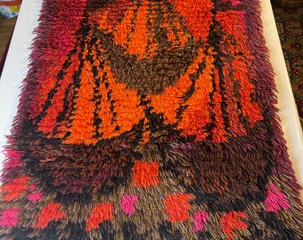 May include: A rectangular, shaggy rug with a vibrant, abstract design. The rug features a color palette of orange, brown, red, and black. The design includes geometric and floral patterns, creating a textured and visually appealing piece.