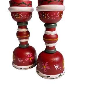 May include: A pair of red wooden candle holders with white and green painted details. The holders feature a floral design with pink and yellow flowers.