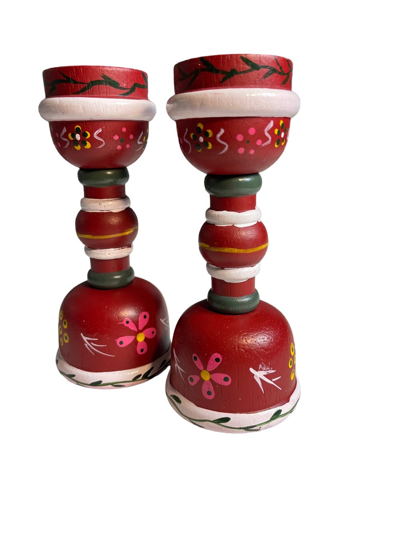 May include: A pair of red wooden candle holders with white and green painted details. The holders feature a floral design with pink and yellow flowers.