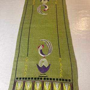 May include: Green embroidered table runner with black vertical lines and decorative floral and rooster designs. The runner has the words "Glad" and "Pask" embroidered on it. The design includes purple, yellow, and white accents.