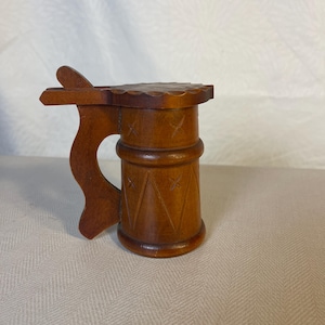 Può includere: Un piccolo boccale di birra in legno con un coperchio a cerniera e un manico intagliato. Il boccale è decorato con un motivo geometrico.
