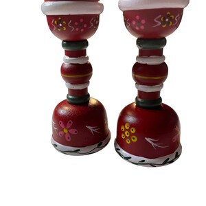 May include: A pair of red wooden candle holders with white and green painted details. The holders feature a floral design with pink, yellow, and black accents.