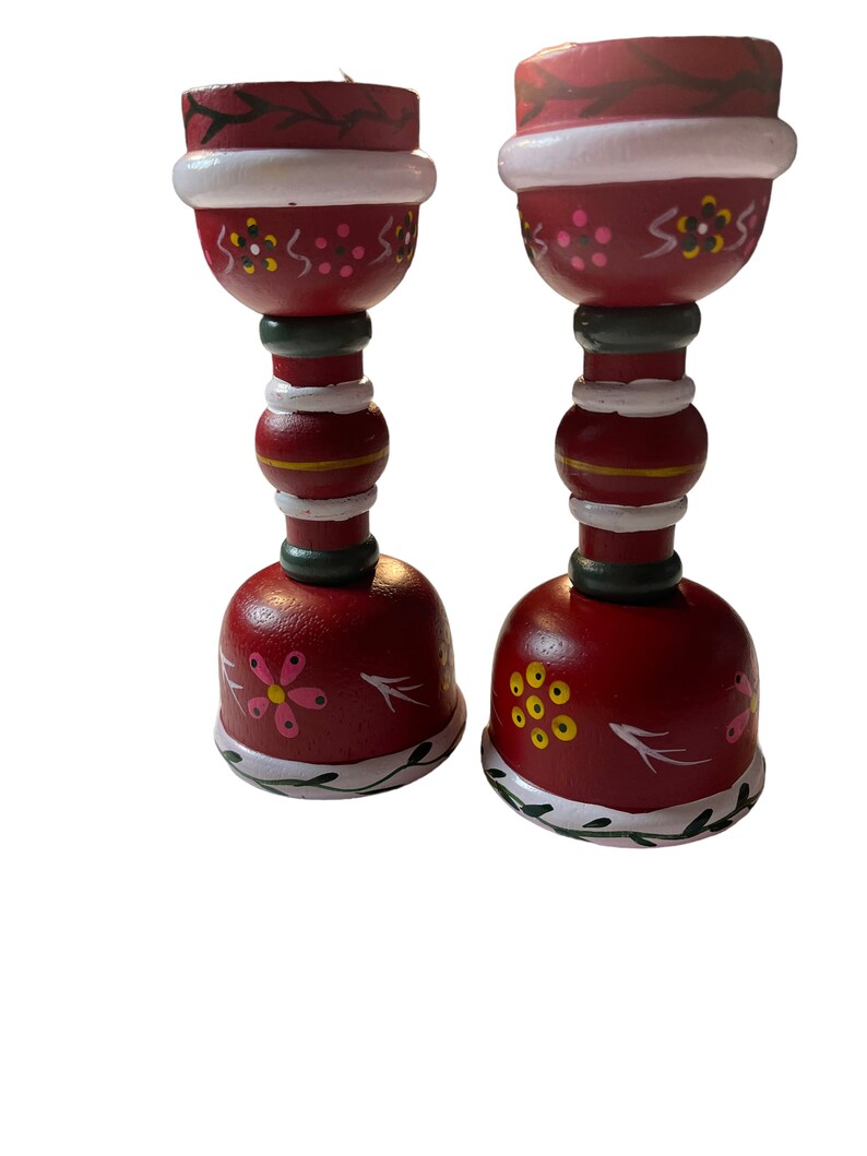 May include: A pair of red wooden candle holders with white and green painted details. The holders feature a floral design with pink, yellow, and black accents.