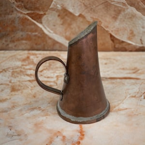 Könnte beinhalten: Ein kleiner Kupferkrug mit Henkel. Der Krug hat ein abgenutztes, altes Aussehen und ist leicht verbeult.