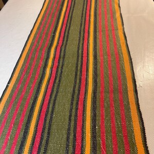 May include: A long, rectangular table runner featuring a woven texture with vertical stripes in olive green, bright red, golden yellow, and thin black lines. The fabric has a consistent, tight weave pattern throughout the entire length of the runner.