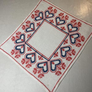 May include: Square white embroidered cloth with a central square and a border of hearts and floral designs in red and navy blue. The cloth is likely handmade and has a vintage aesthetic.