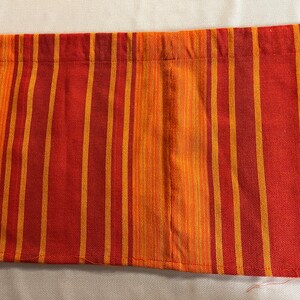May include: Close-up of a fabric with vertical stripes in shades of orange and red. The stripes alternate between a darker red-orange and a lighter, golden orange. The fabric appears to be woven, with a visible texture.