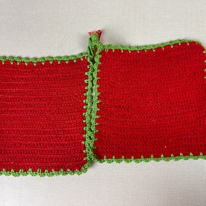 Puede incluir: Dos agarraderas rojas de ganchillo con un borde verde festoneado. Las agarraderas son de forma cuadrada y están conectadas en la parte superior con un lazo verde para colgar. El trabajo de ganchillo es visible.