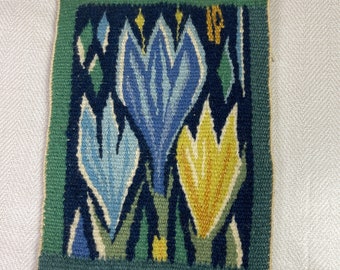 May include: A square, woven tapestry featuring a floral design. The central image showcases three stylized flowers in shades of blue and yellow against a dark blue background. The border is green, and the piece is set against a light-colored, textured backdrop.
