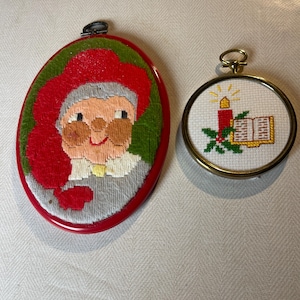 May include: Two vintage Christmas ornaments. One is oval-shaped with a red border, featuring a stitched image of a Santa Claus-like figure. The other is round, with a gold-colored frame, depicting a candle, book, and holly.